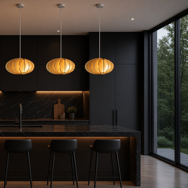 A modern kitchen interior with five warm glowing pendant lights hanging above a dark island, black cabinetry, bar stools, and large windows showing greenery outside.