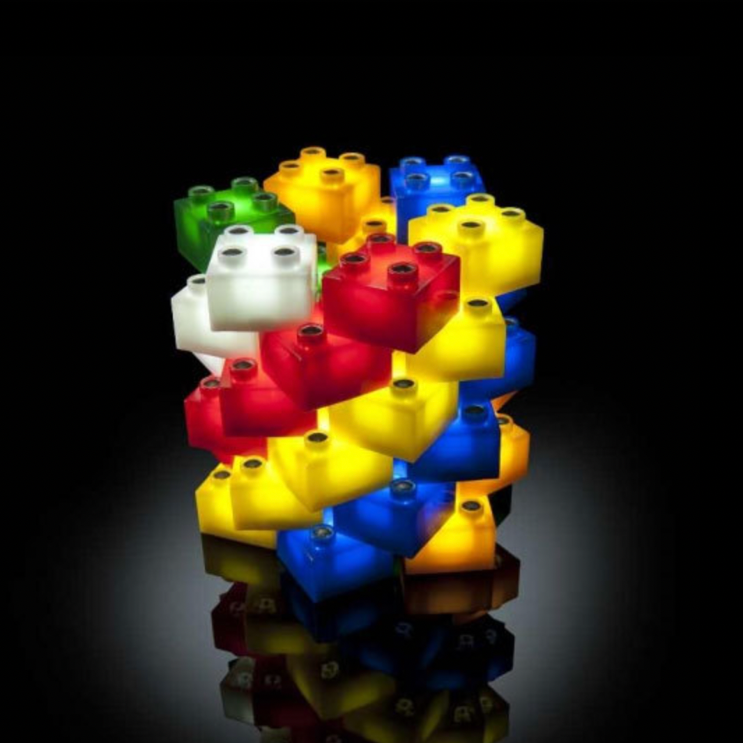 A cluster of translucent toy building-block-style lights in red, yellow, blue, green, and white, arranged in a tower against a black background with a faint reflection below.
