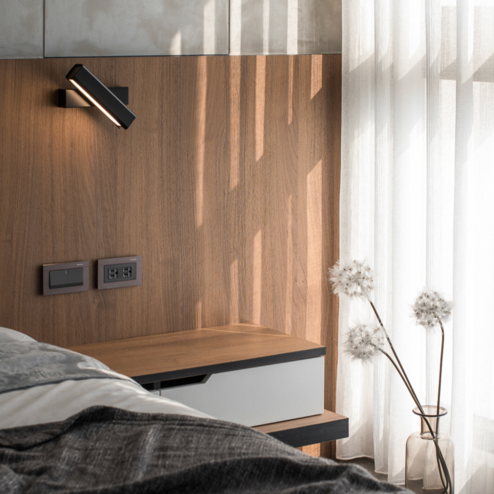 A modern black LED wall light with a swivel head, mounted on a wall above a round wooden table, with a striped pillow on the table and a book on the floor.